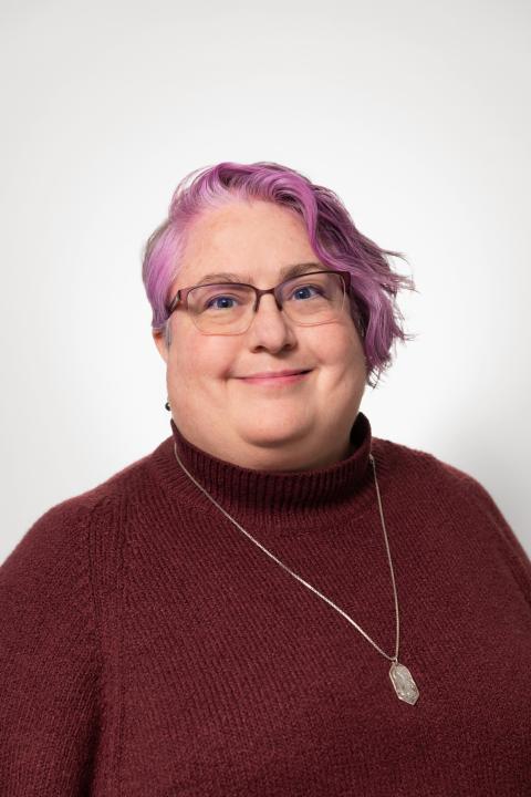 A white woman with short lavendar colored hair. She is wearing a burgundy sweater and a silver necklace.